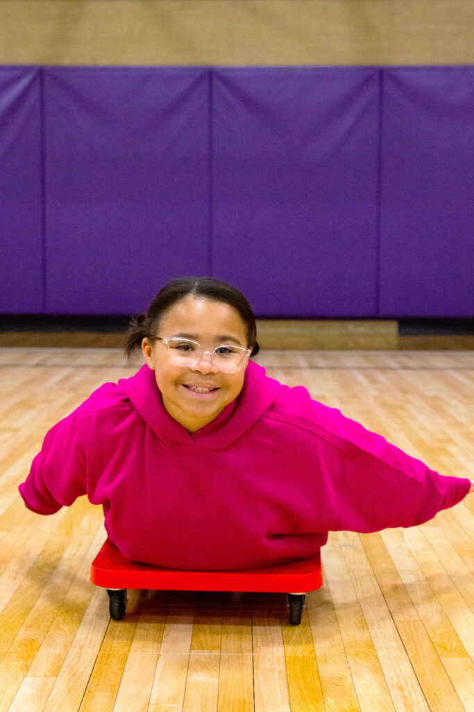 School age girl sliding on a wheelie cart in the gym